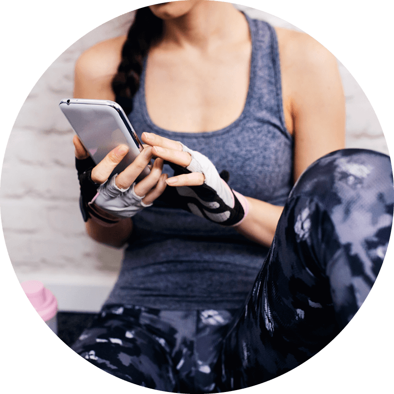 Eversense user holding smartphone, wearing workout attire and fingerless gloves, sitting indoors against a white brick wall.